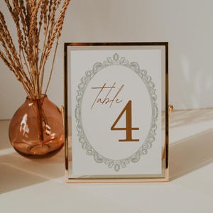 Può includere: Un segnaposto con cornice dorata con il numero 4 in oro. La carta ha un bordo ovale e ornato e la parola "Table". Un vaso con piante secche è sullo sfondo.