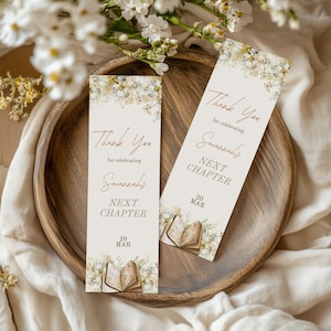 May include: Two rectangular bookmarks with floral designs and the text "Thank You for celebrating Savannah's Next Chapter 20 MAR". The bookmarks are on a wooden tray with white flowers.