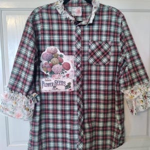 May include: A long-sleeved plaid shirt in red, green, and white. The shirt features a floral patch with the words "Choice Flower Seeds." The cuffs and collar have a ruffled floral trim. A pocket is on the left chest.