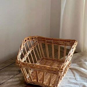 May include: A light brown woven wicker basket with an open-top design. The basket features a rectangular base with vertical slats forming the sides and a reinforced rim. The natural wood material displays a textured, hand-woven construction.