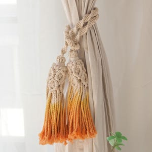 May include: Cream-colored macrame curtain tiebacks with ombre orange tassels. The tiebacks are knotted and twisted, holding back beige curtains. A small green plant is visible in the lower right corner.
