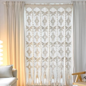 May include: Cream-colored macrame curtain with geometric and butterfly patterns. The curtain hangs in front of a window, with white curtains on either side. The text "LIBRE VOYAGE" is visible at the bottom right.