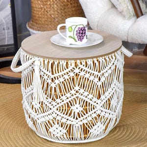 May include: A round macrame side table with a light wood top. The white macrame has a woven pattern and a tassel. A white mug with a grape design sits on a white saucer on the table. The text "LIBRE VOYAGE" is visible.