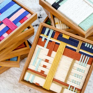 K&ouml;nnte beinhalten: Eine Sammlung quadratischer Holztabletts mit geflochtenen Seileins&auml;tzen in verschiedenen Farbmustern, darunter Blau, Rosa, Rot, Gelb und Creme. Der Text LIBRE VOYAGE ist in der unteren rechten Ecke des Bildes zu sehen.