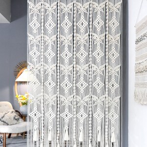 Puede incluir: Cortina de macramé color crema con un patrón geométrico. La cortina presenta formas de diamantes y mariposas, con borlas en la parte inferior. El texto "LIBRE VOYAGE" es visible en la parte inferior derecha.