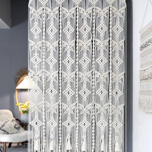 May include: A large off-white macrame curtain featuring intricate knotted patterns, including repeating diamond shapes and vertical twisted cord columns. The bottom edge is finished with a series of hanging tassels, and the top is suspended from a rod.