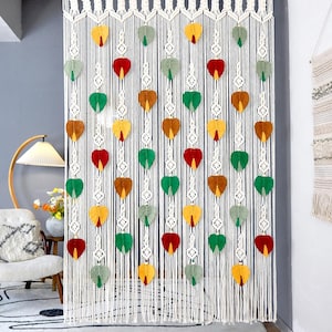 May include: A macrame curtain with a white woven design and hanging strands. Colorful, heart-shaped accents in red, green, yellow, and brown are evenly spaced throughout the curtain. The text "LIBRE VOYAGE" is visible at the bottom.