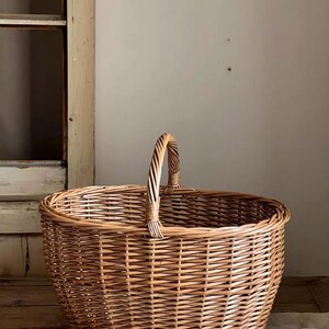 May include: A round, woven wicker basket featuring a natural light brown finish. The basket has a sturdy, arched handle attached to the center of the rim, with a consistent, tight weave pattern throughout the body and a reinforced circular base.