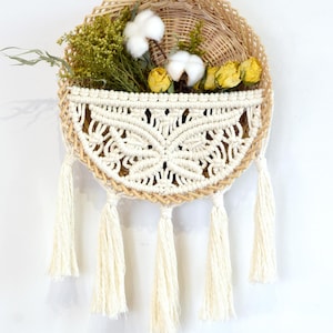 May include: A circular woven wall hanging with a macrame pocket, filled with dried flowers and cotton. The macrame is cream-coloured with a detailed pattern, and the hanging has cream-coloured tassels. The basket contains yellow roses and green foliage.