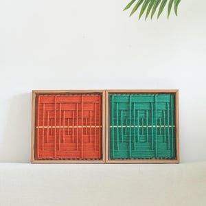 K&ouml;nnte beinhalten: Zwei quadratische Wandbilder in Holzrahmen mit gewebten Garnmustern. Ein Bild zeigt ein leuchtend orangefarbenes Muster, das andere ein passendes gr&uuml;nes Muster. Beide weisen eine horizontale Linie und den Text LIBRE VOYAGE auf.