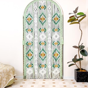 May include: A macrame door curtain with a green, white, and gold geometric design. The curtain is arched at the top and has a fringe at the bottom. The text "LIBRE VOYAGE" is at the bottom right.
