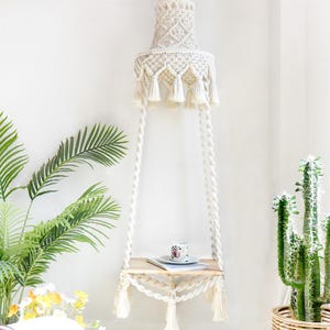 May include: A macrame hanging shelf with a natural wood shelf and cream-colored macrame detailing. The shelf is suspended by twisted rope and features decorative tassels. A matching macrame lampshade hangs above. The text "LIBRE VOYAGE" is visible at the bottom.