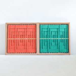Könnte beinhalten: Zwei quadratische Wandbilder in Holzrahmen mit geflochtenen Seilmustern. Ein Bild ist rot mit einem horizontalen gelben Faden, das andere ist grün mit einem horizontalen gelben Faden. Beide zeigen ein geometrisches Gittermuster.