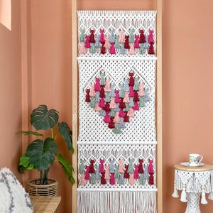 May include: A macrame wall hanging with a wooden frame. The white macrame features a heart design made of colorful tassels in shades of pink, red, and gray. The bottom has a fringe. The text "LIBRE VOYAGE" is at the bottom.
