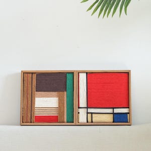 K&ouml;nnte beinhalten: Zwei quadratische Holzrahmen mit geometrischer Wandkunst aus fest gewickeltem Garn. Das linke St&uuml;ck zeigt braune, beige, wei&szlig;e, gr&uuml;ne und rote Streifen. Das rechte St&uuml;ck zeigt ein Gitter aus roten, wei&szlig;en, beigen, blauen und schwarzen Garnlinien.