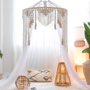 May include: A cream macrame canopy with a sheer white curtain. The scene includes a woven lantern, a round candle holder, a decorative pillow, and a rug, creating a bohemian aesthetic.