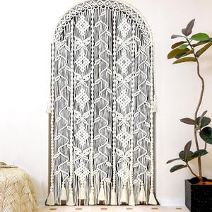 Pode incluir: Uma tapeçaria de macramé creme com uma parte superior arqueada. O design intrincado apresenta padrões geométricos e borlas na parte inferior. O texto "LIBRE VOYAGE" é visível no canto inferior direito. Uma planta em vaso está à direita.