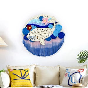 May include: A circular wall hanging with a crocheted whale design, incorporating blue, pink, and white yarn. The artwork includes floral details and a fringed bottom. It's displayed above a sofa with decorative cushions, creating a coastal theme.