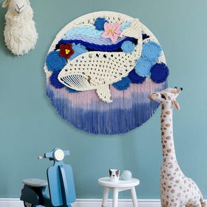 May include: A circular macrame wall hanging featuring a cream-colored whale silhouette surrounded by blue and light blue yarn swirls, a pink flower, and a dark red flower, with a gradient fringe transitioning from light pink to deep blue.