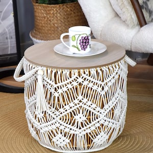 Può includere: Un tavolino rotondo con piano in legno chiaro e base cilindrica rivestita in corda macramè bianca su una struttura in metallo dorato. Il tavolo presenta due maniglie laterali con nappe bianche e sostiene una tazza in ceramica bianca.