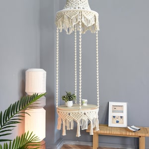 Può includere: Una mensola sospesa in macramè color crema con un piano rotondo in legno, sospesa da cordoncini intrecciati. La mensola è adornata con una piccola pianta e una tazza. Il design include una tenda in macramè abbinata. Il testo "LIBRE VOYAGE" è visibile.