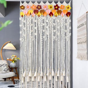 Pode incluir: Cortina de macramê creme com um design floral e de folhas. A parte superior apresenta flores de crochê em tons de rosa, amarelo, laranja e vermelho. A cortina tem fios verticais longos com borlas na parte inferior. O texto "LIBRE VOYAGE" é visível.