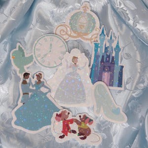 Könnte beinhalten: Eine Sammlung glitzernder Aufkleber mit Cinderella-Motiven. Enthält Bilder von Cinderella im Hochzeitskleid, einem Glasschuh, einer Kutsche, einem Schloss und anderen Charakteren. Die Aufkleber sind vor einem hellblauen, strukturierten Hintergrund platziert.