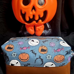 May include: An orange plastic jack-o'-lantern sits above a Halloween-themed cosmetic bag. The bag features a blue fabric top with pumpkin, candy, and bat patterns, and a brown leather bottom. The background is black.