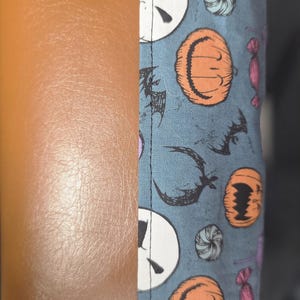 May include: A brown leather and patterned fabric item. The fabric features Jack Skellington faces, orange pumpkins, bats, and candies on a blue background. The item is likely a case or cover.