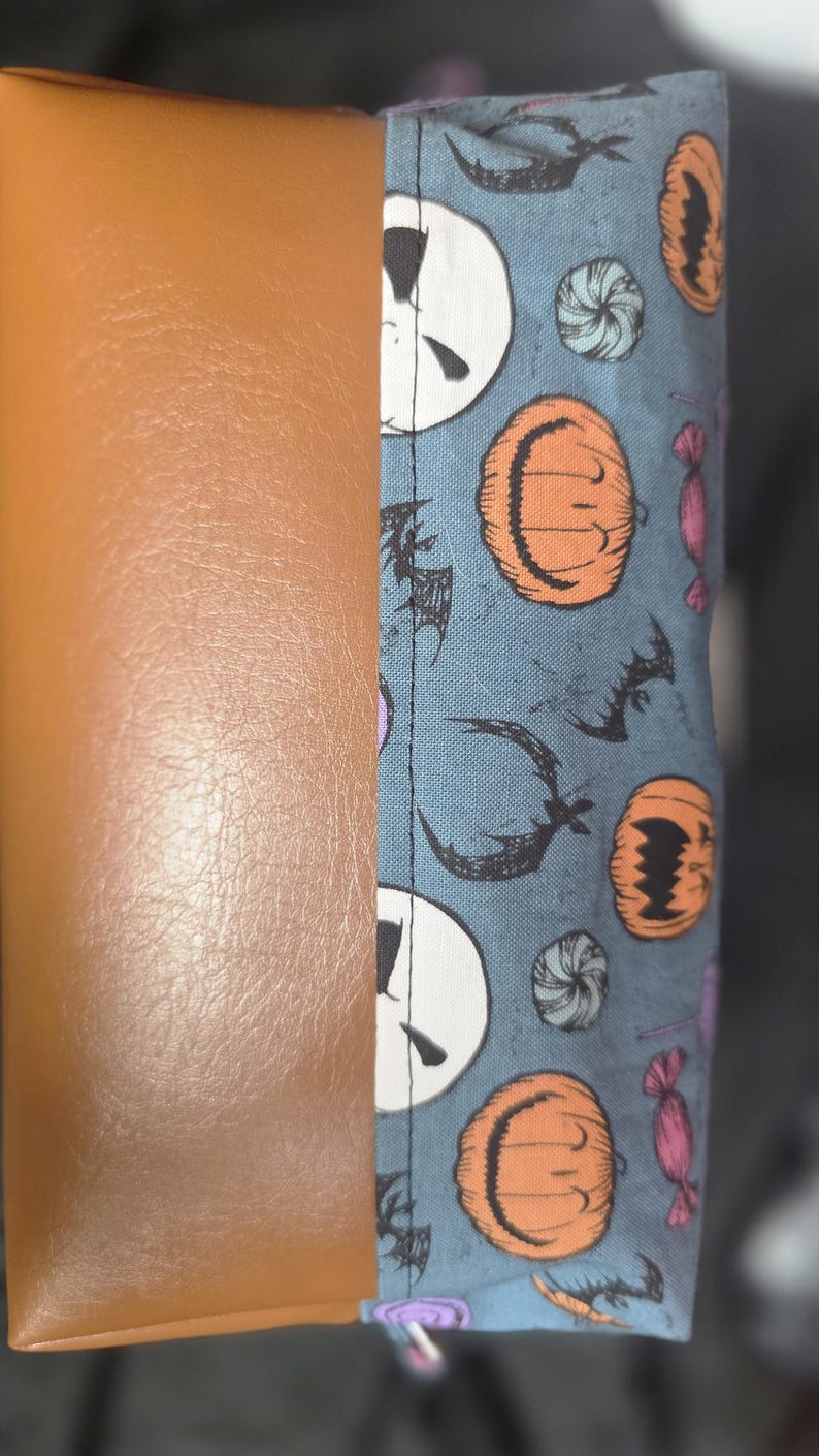 May include: A brown leather and patterned fabric item. The fabric features Jack Skellington faces, orange pumpkins, bats, and candies on a blue background. The item is likely a case or cover.