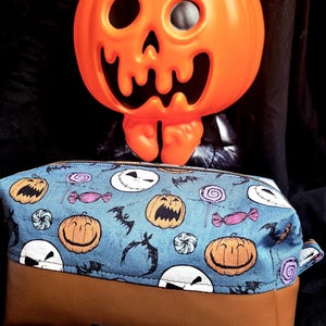 May include: A Halloween-themed pencil case with a blue fabric top featuring jack-o'-lanterns, bats, and candy. The bottom is brown. An orange plastic jack-o'-lantern decoration is in the background.