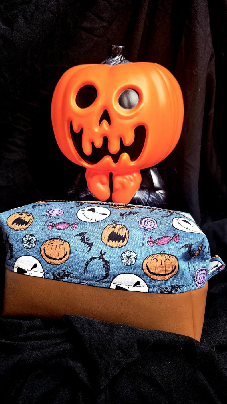 May include: A Halloween-themed pencil case with a blue fabric top featuring jack-o'-lanterns, bats, and candy. The bottom is brown. An orange plastic jack-o'-lantern decoration is in the background.