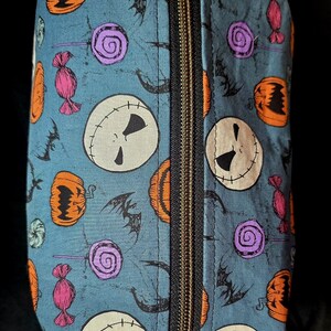 May include: A rectangular, zippered cosmetic bag with a teal background. The bag is patterned with Jack Skellington faces, orange pumpkins, purple lollipops, pink wrapped candies, and black bats. The zipper is black.