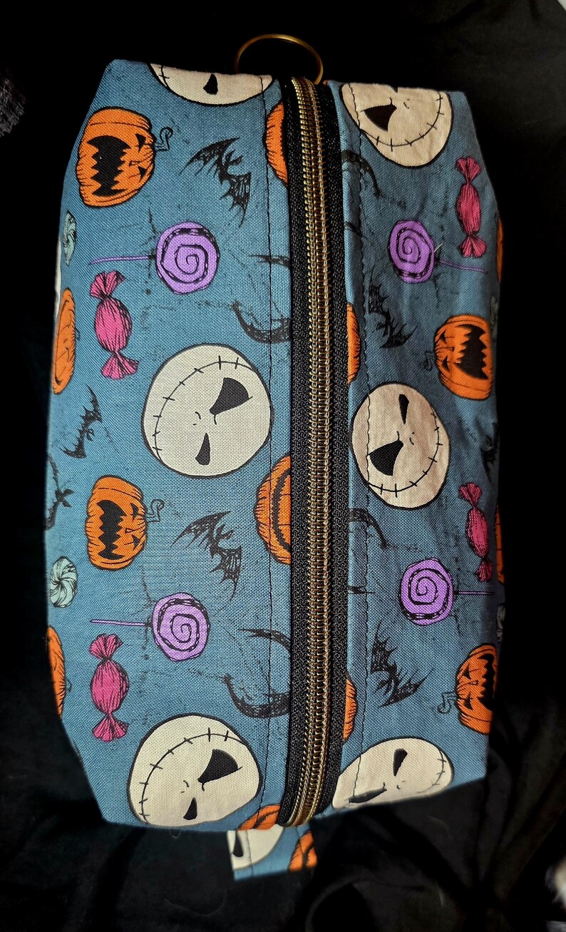 May include: A rectangular, zippered cosmetic bag with a teal background. The bag is patterned with Jack Skellington faces, orange pumpkins, purple lollipops, pink wrapped candies, and black bats. The zipper is black.