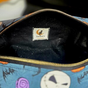May include: Open blue cosmetic bag with a gold zipper, featuring a pattern of Jack Skellington faces, bats, and Halloween candies. The interior is black with a small tag that reads "Torina's".