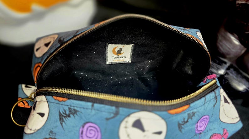 May include: Open blue cosmetic bag with a gold zipper, featuring a pattern of Jack Skellington faces, bats, and Halloween candies. The interior is black with a small tag that reads "Torina's".