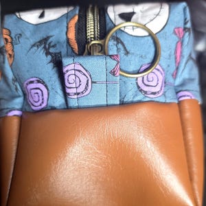 May include: A small, square pouch with a brown leather bottom and a blue fabric top featuring a Jack Skellington pattern. The pouch has a gold zipper and a gold ring attached. The fabric design includes purple lollipops and Halloween-themed elements.