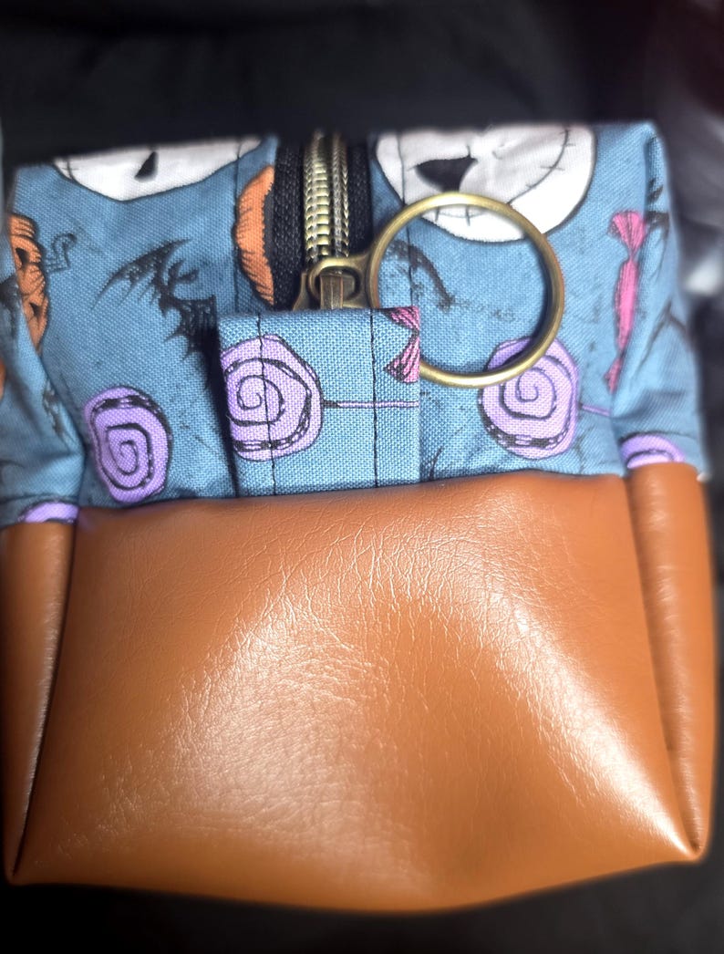 May include: A small, square pouch with a brown leather bottom and a blue fabric top featuring a Jack Skellington pattern. The pouch has a gold zipper and a gold ring attached. The fabric design includes purple lollipops and Halloween-themed elements.