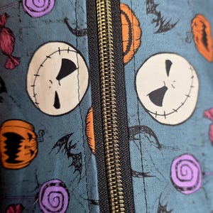 May include: Close-up of a teal fabric with a repeating pattern of Jack Skellington faces, orange pumpkins, purple swirl candies, and black bats. A gold zipper runs down the center of the image.
