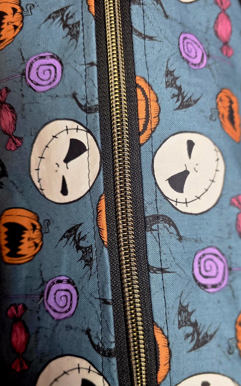 May include: Close-up of a teal fabric with a repeating pattern of Jack Skellington faces, orange pumpkins, purple swirl candies, and black bats. A gold zipper runs down the center of the image.