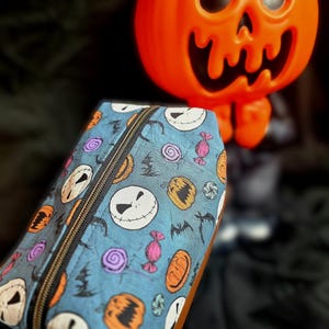 May include: A blue pencil case with a black zipper and a gold ring pull. The case is patterned with Jack Skellington faces, pumpkins, bats, and candy. An orange pumpkin decoration is in the background.