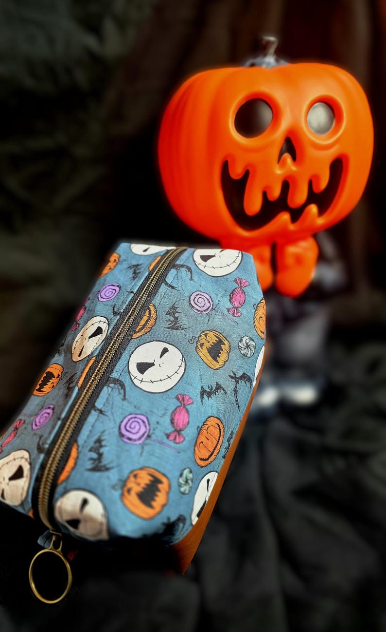 May include: A blue pencil case with a black zipper and a gold ring pull. The case is patterned with Jack Skellington faces, pumpkins, bats, and candy. An orange pumpkin decoration is in the background.