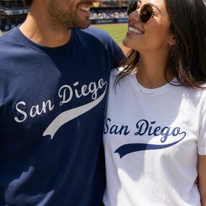 May include: A navy blue t-shirt with white San Diego text and a white t-shirt with navy blue San Diego text. The navy blue shirt features a white swoosh graphic under the text, while the white shirt features a navy blue swoosh graphic under the text.
