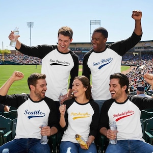 May include: Five people wearing white baseball-style shirts with black three-quarter sleeves. Each shirt features a city name in a script font with a swoosh: San Francisco, San Diego, Seattle, Pittsburgh, and Philadelphia. They are in a stadium.