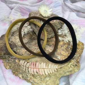 Könnte beinhalten: Drei kreisförmige Haarbänder in den Unifarben Senfgelb, Dunkelbraun und Schwarz. Jedes Band weist eine glatte, matte Stofftextur und ein schlichtes, durchgehendes Ringdesign auf, präsentiert als ein Set aus drei verschiedenen Haaraccessoires.