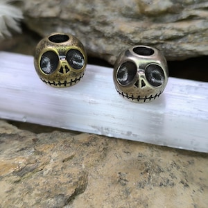 Könnte beinhalten: Zwei kugelförmige Metallperlen mit einem geschnitzten Totenkopfgesicht, großen hohlen Augenhöhlen, einer kleinen dreieckigen Nase und einem genähten Mund. Eine Perle hat ein dunkles Messing-Finish, die andere ein poliertes, silberfarbenes Finish.