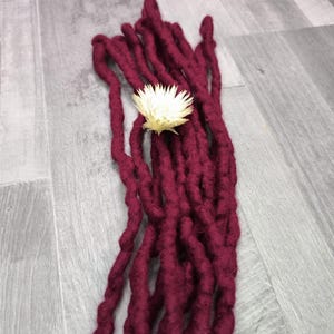 Puede incluir: Un conjunto de rastas sintéticas largas de color burdeos intenso con una apariencia texturizada y retorcida. Una pequeña flor seca de color crema está colocada cerca del centro superior del manojo, descansando sobre las fibras rojas.