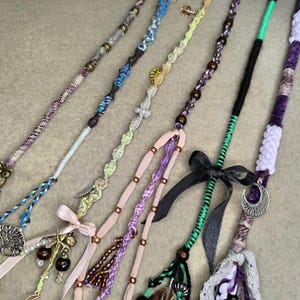 May include: An assortment of handmade hair wraps in various colors and designs. Each wrap features unique embellishments like beads, ribbons, and charms. The color palette includes blues, purples, greens, and pinks, with decorative bows and metallic accents.