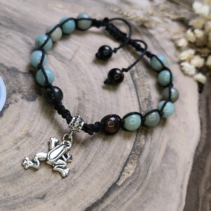 May include: A beaded bracelet featuring a frog charm. The bracelet has a black cord with light blue and black beads. The frog charm is silver-colored. The bracelet is adjustable.