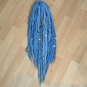 Könnte beinhalten: Ein Bündel hellblauer Dreadlocks mit verschiedenen dekorativen Perlen und Akzenten. Die Dreadlocks sind vertikal angeordnet, mit einer Mischung aus Texturen und Blautönen. Einige Dreadlocks haben weiße und Holzperlen.